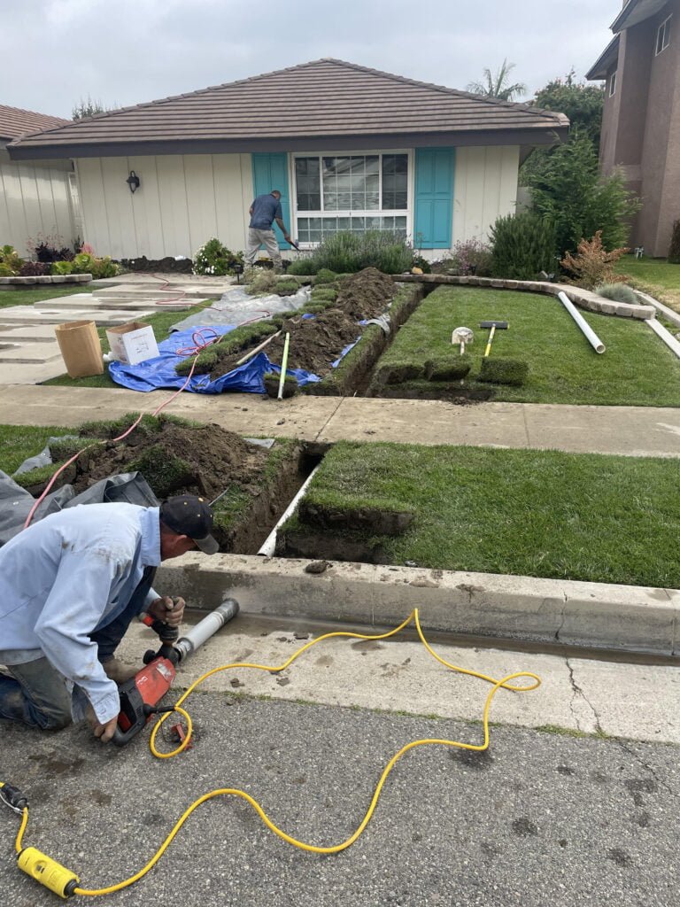Technician drilling a hole through the street curb for drainage installation, while another technician digs a trench in the lawn.