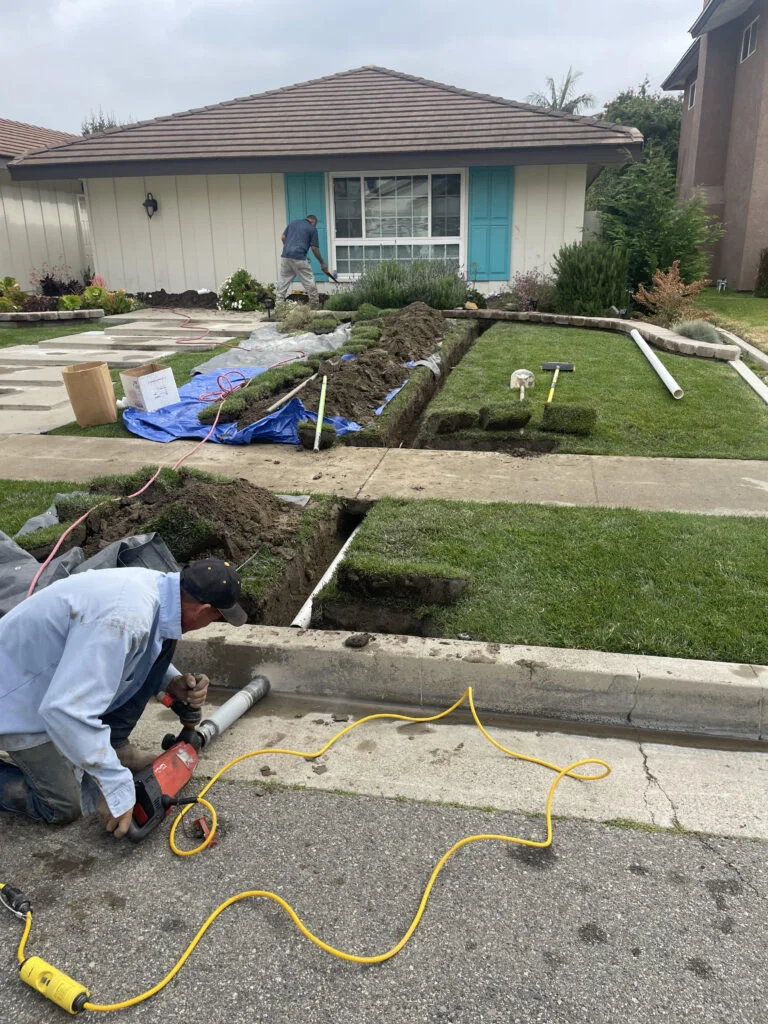 Technician drilling a hole through the street curb for drainage installation, while another technician digs a trench in the lawn.